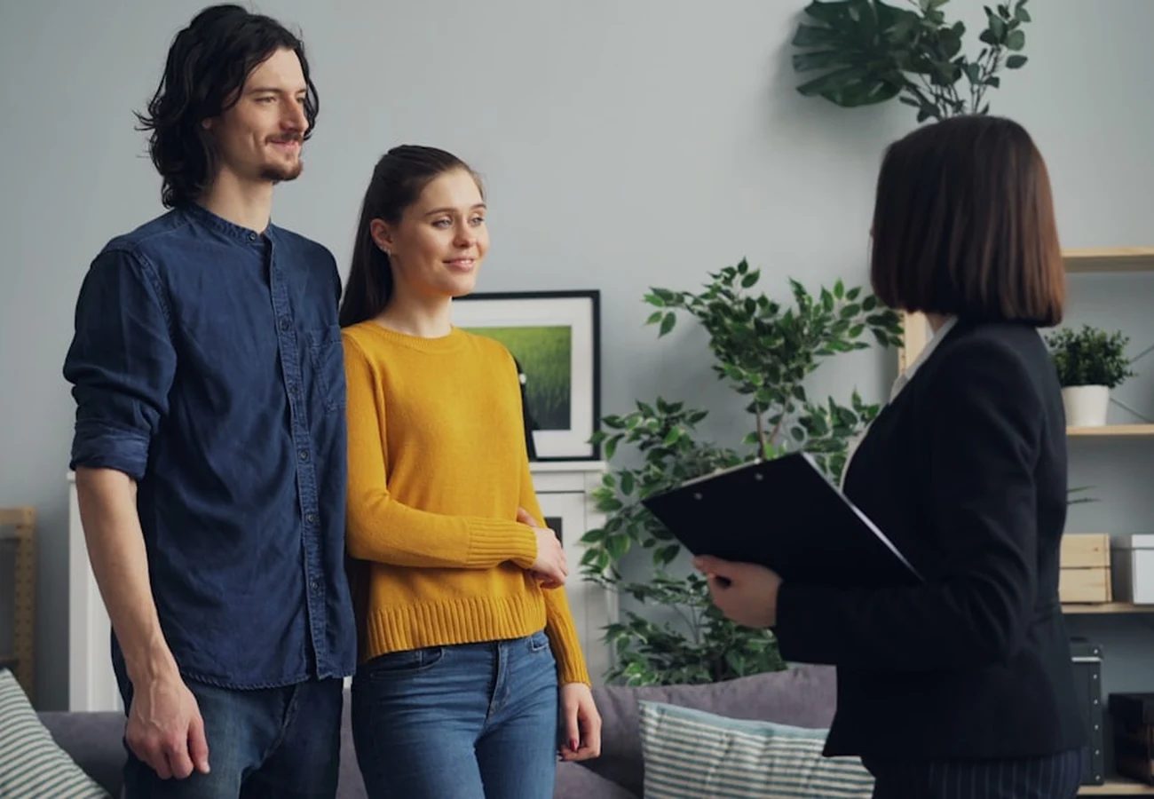 A couple talking with an agent who holds a clipboard in a bright living room