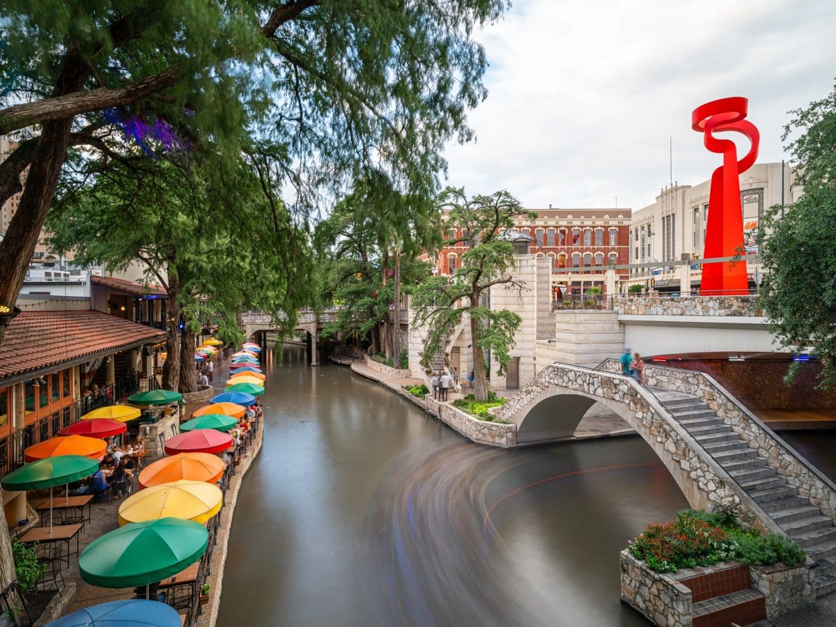 The San Antonio River Walk, with cafe umbrellas along the water and an arched stone footbridge