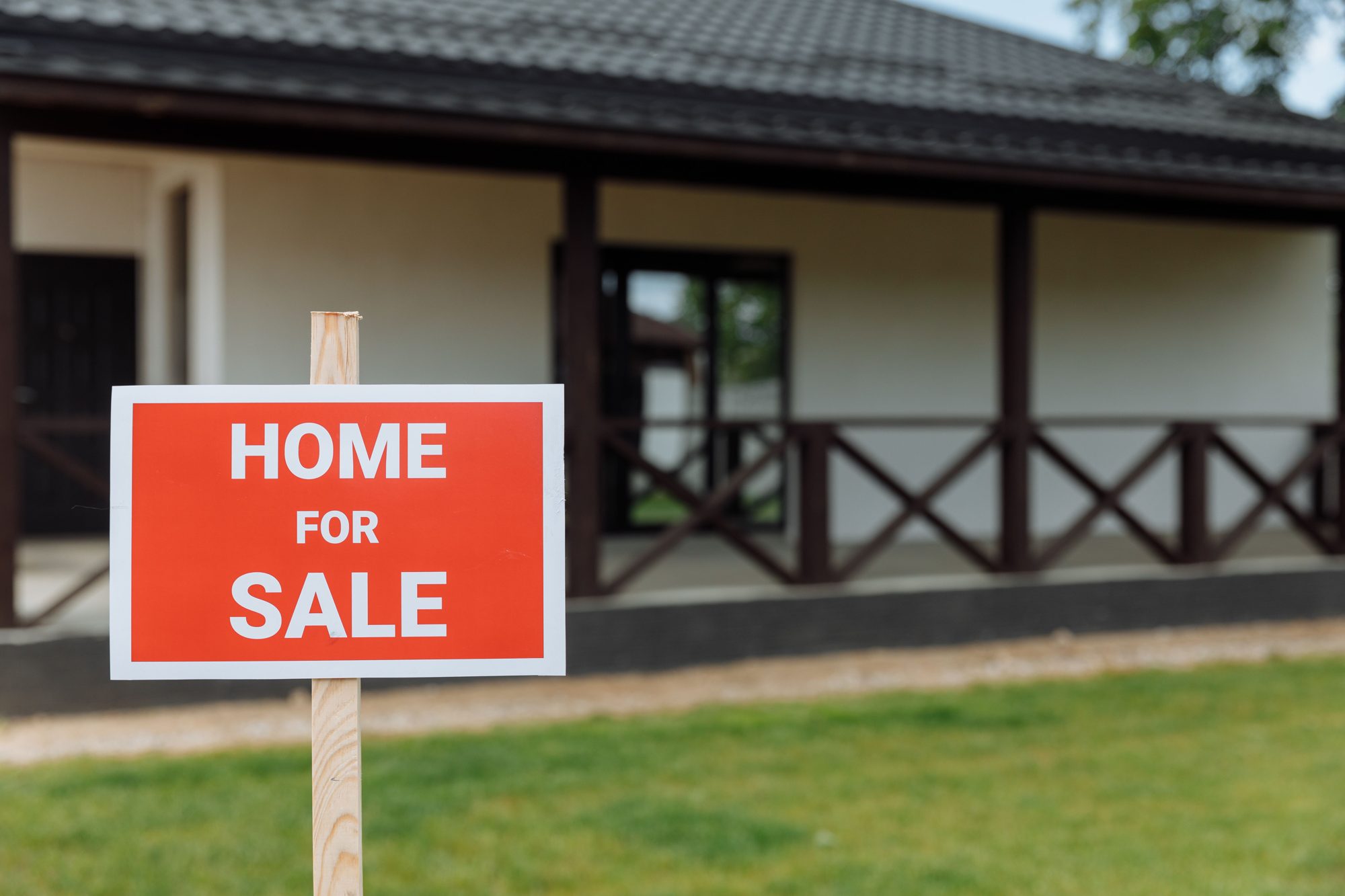 A red Home For Sale sign on the lawn in front of a house with a covered porch