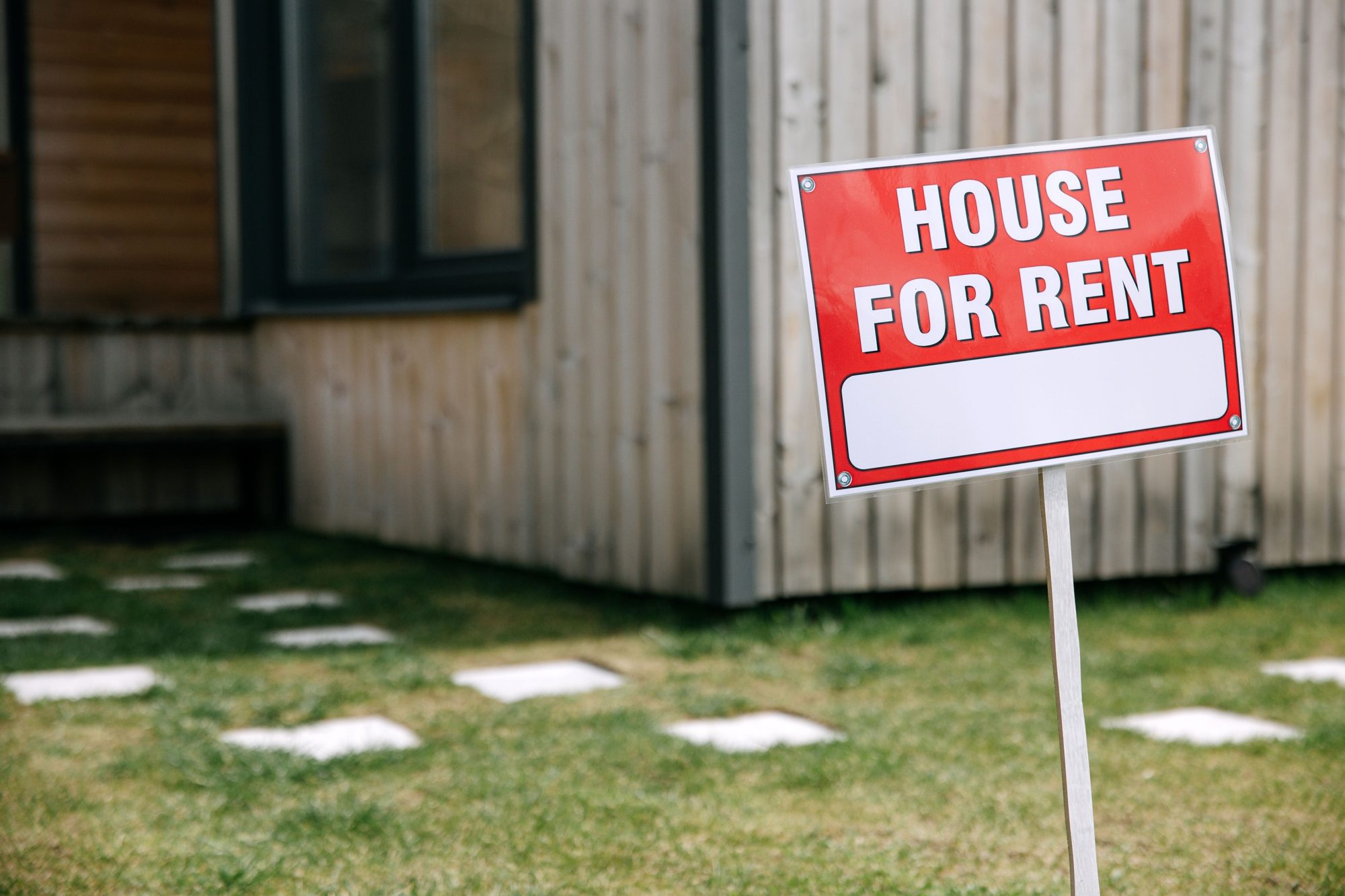 A red House For Rent sign on the lawn beside a wood-clad house