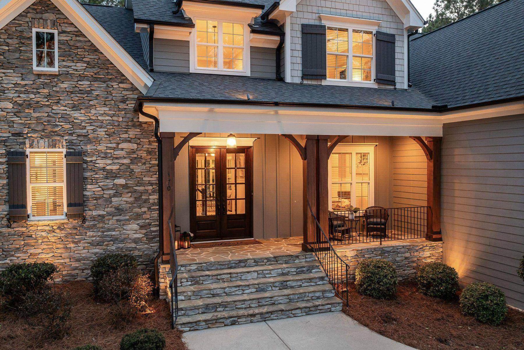 A stone-and-siding craftsman home with its front porch lights on at dusk