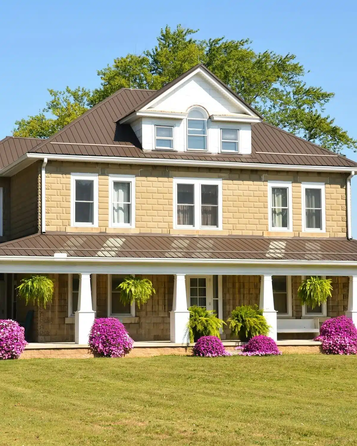 A two-story Texas farmhouse with a brown metal roof and flowering front beds
