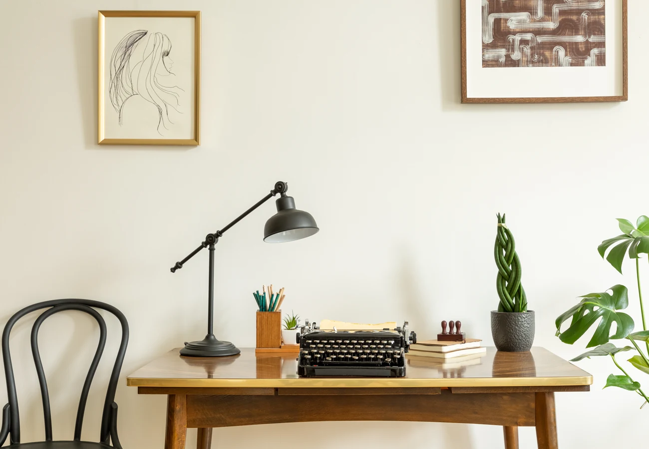 A writing desk by a window with a typewriter, a lamp and a potted plant