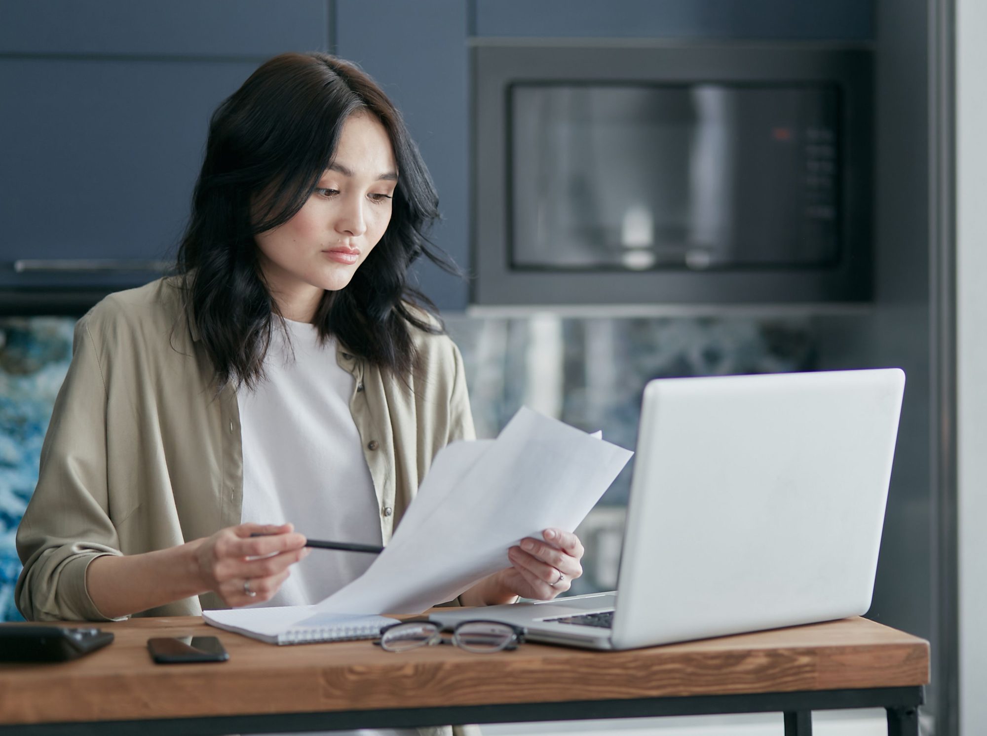 A woman reading through paperwork at a desk beside her laptop