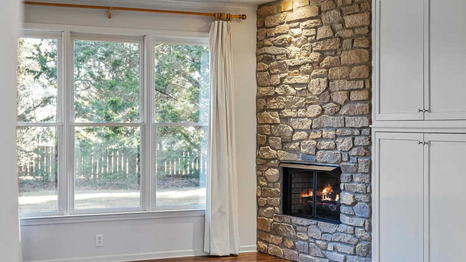 An empty living room with a stone fireplace, wood floors and tall windows