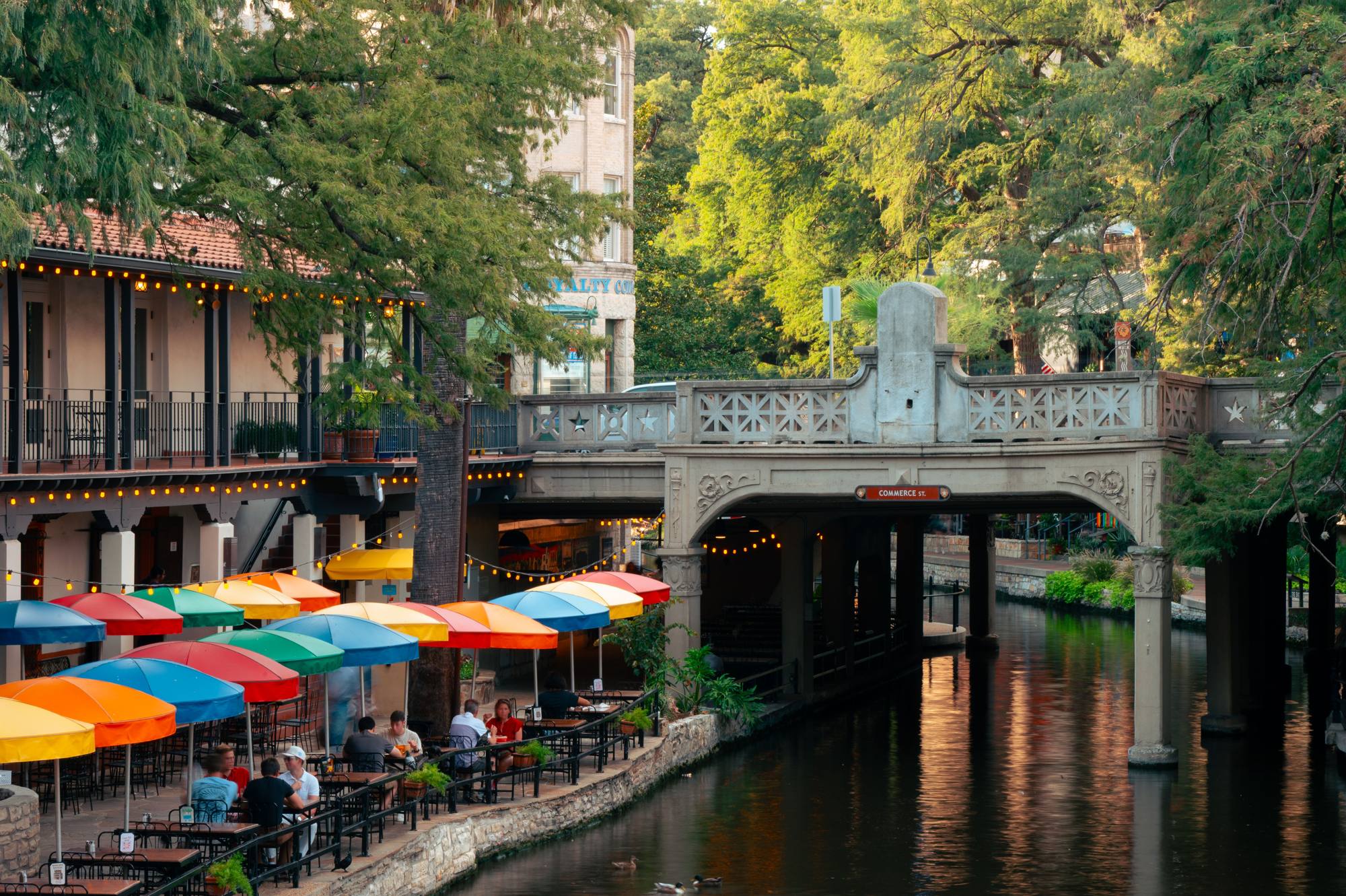 Bright market umbrellas along the San Antonio River Walk beside a stone footbridge